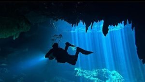 A diver silhouetted against the blue glow of sunlight beams in a cenote cave.
